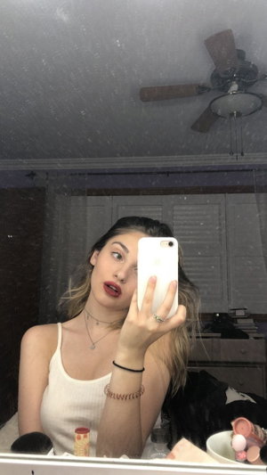 Woman in white tank top taking mirror selfie in bedroom.