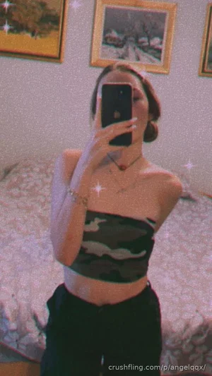 Woman in camo crop top and black pants takes mirror selfie in bedroom.