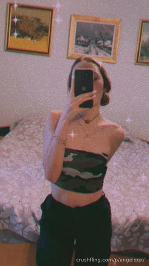 Woman in camo crop top and black pants takes mirror selfie in bedroom.