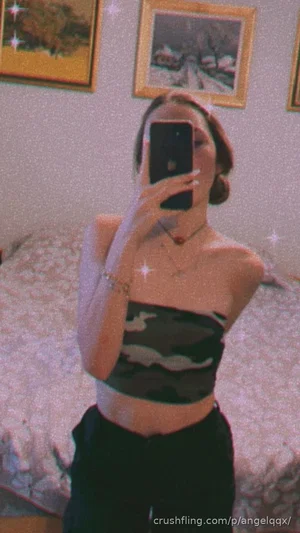Woman in camo crop top and black pants takes mirror selfie in bedroom.