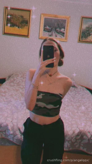 Woman in camo crop top and black pants takes mirror selfie in bedroom.