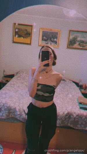 Woman in camo crop top and black pants takes mirror selfie in bedroom.
