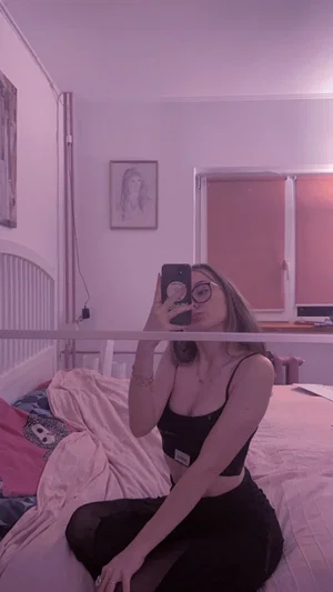 Woman in black crop top and pants sits on bed, taking mirror selfie in bedroom.