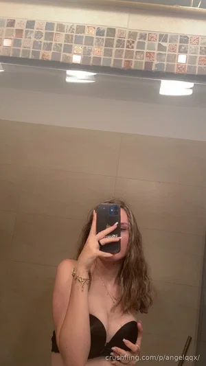 Woman with wavy hair in black bra taking mirror selfie in tiled bathroom.
