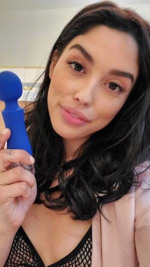 Brunette in fishnet top and pink blazer holds blue toy indoors.
