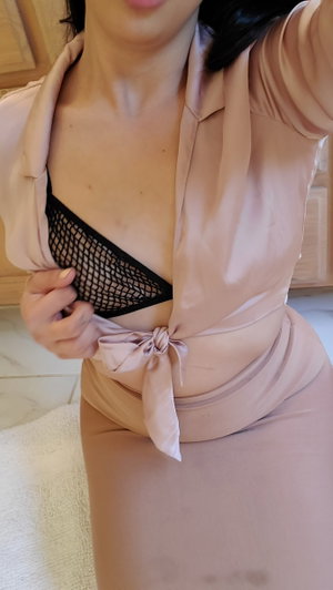 Woman with dark hair in open pink blouse over black mesh bra in bathroom.