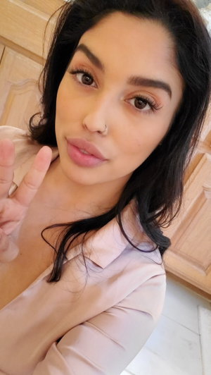 Curvy woman with dark hair in sheer pink top, peace sign indoors.