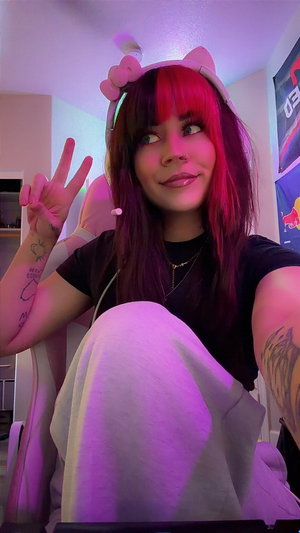 Woman with purple hair and cat ear headband, wearing black shirt and white pants, making peace sign