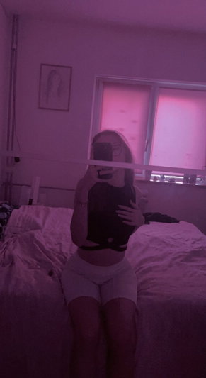 Woman in black crop top and white shorts sits on bed in pink-lit room, holding phone for mirror