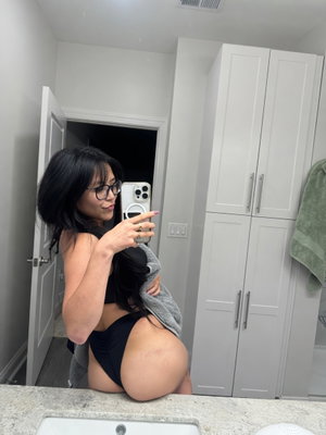 Woman with black hair and glasses in black underwear and towel, taking mirror selfie in bathroom.