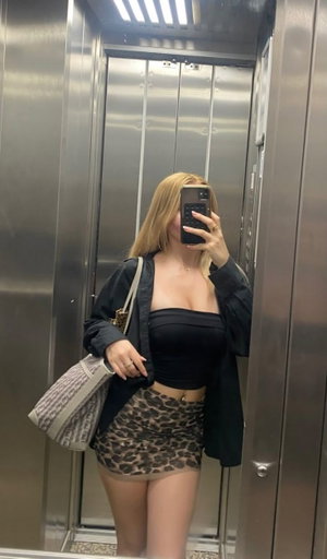 Woman with blonde hair in elevator, wearing black crop top, leopard print skirt, and black jacket,