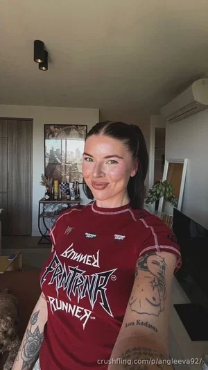Woman with tattoos wearing red graphic tee, taking selfie indoors in living room.