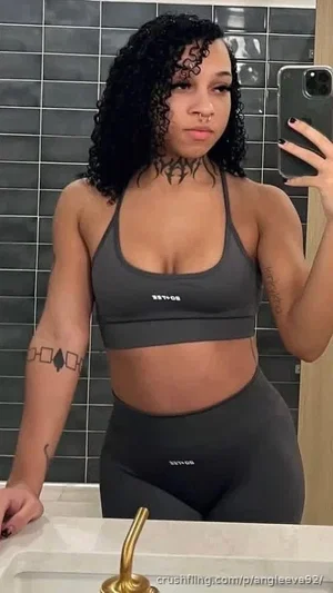 Woman with curly hair in gray sports bra and leggings, taking mirror selfie in bathroom.