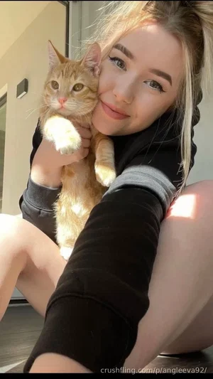 Woman with blonde hair holding a ginger cat, wearing black sweatshirt, sitting indoors.