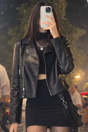 Slim woman in leather jacket, crop top, mini skirt, fishnets outdoors.