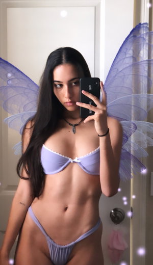 Brunette in purple bikini with fairy wings takes bathroom mirror selfie.