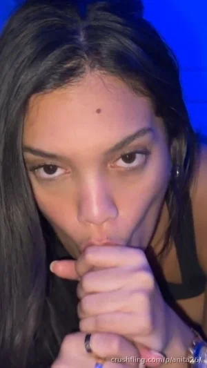 Woman with dark hair and black top, holding hand near mouth, blue background.