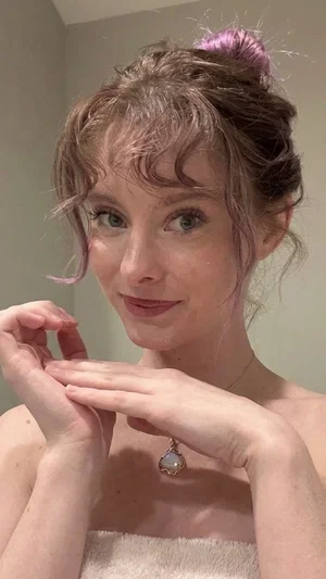 Woman with pink-tipped hair, wearing a towel, smiling in a mirror selfie indoors.