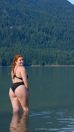 Woman with red hair in black bikini standing in lake, mountains in background.