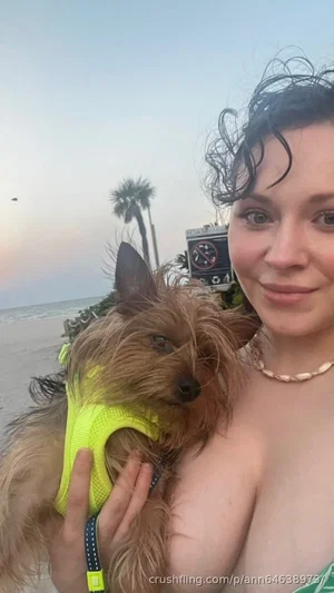 Woman with curly hair holding a small dog in a neon vest on a beach at dusk.