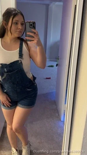 Woman in white tank top and denim overalls taking mirror selfie in indoor hallway.