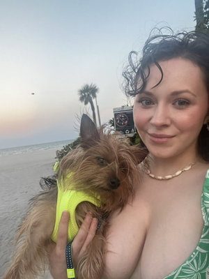 Woman with dark hair holding small dog on beach, wearing green patterned top, bare shoulders.