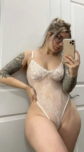 Woman with tattoos wears white lace bodysuit, takes mirror selfie in indoor setting.