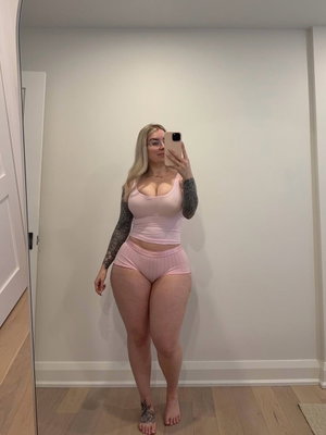 Woman with blonde hair and tattoos wears pink top and shorts, standing in front of mirror indoors.