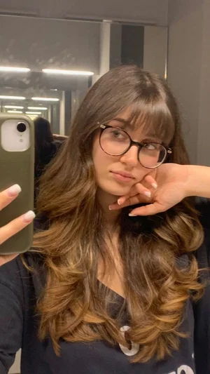 Woman with long brown hair and glasses takes mirror selfie in dark top.