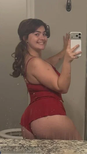 Curvy woman in red dress taking bathroom mirror selfie.