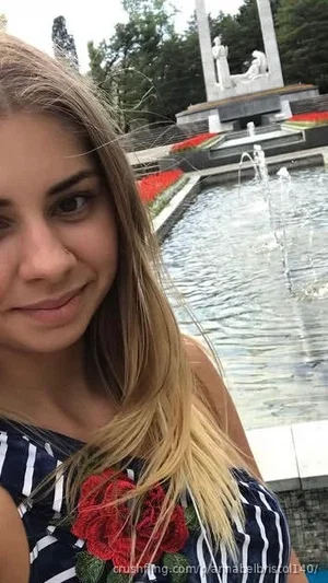 Woman with long blonde hair in striped top with red flower, near fountain and monument.