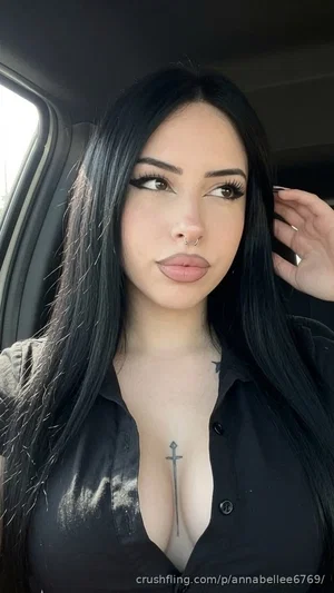 Woman with long black hair and dark makeup in black button-up shirt, sitting in car.