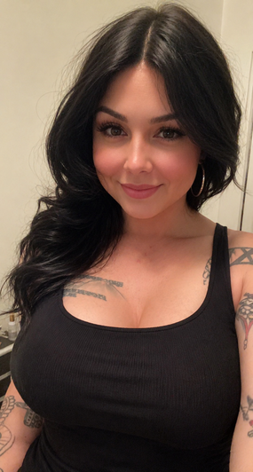 Woman with long black hair and tattoos wearing black tank top indoors.