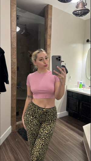 Woman in pink crop top and leopard pants taking mirror selfie in bathroom.
