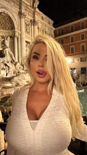 Blonde woman in white dress posing outdoors near a fountain at night.