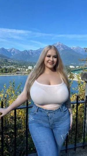 Woman with long blonde hair in white crop top and jeans, standing outdoors near mountains and water.