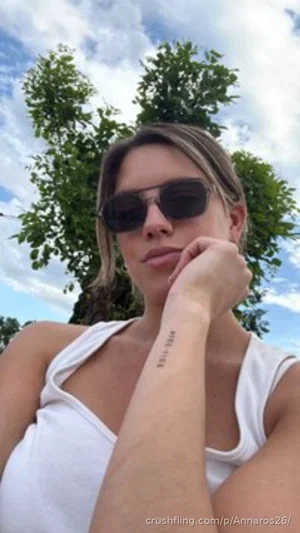 Woman in white tank top and sunglasses outdoors with trees and sky behind her.