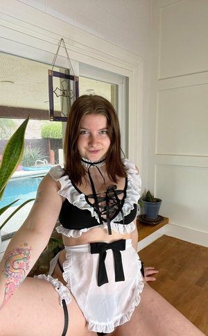 Brunette in black and white maid lingerie posing indoors by window.
