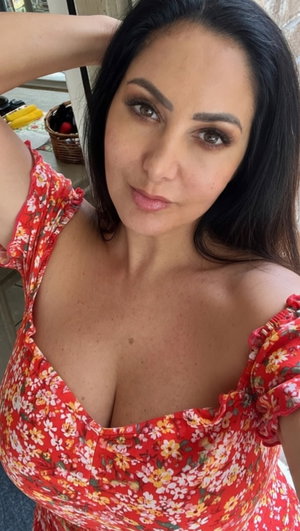 Woman with dark hair in red floral top posing indoors.