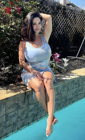 Woman in light blue dress sits by pool, legs dangling in water, hand in hair.