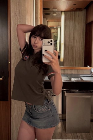 Woman in olive crop top and denim shorts takes mirror selfie in modern bathroom.