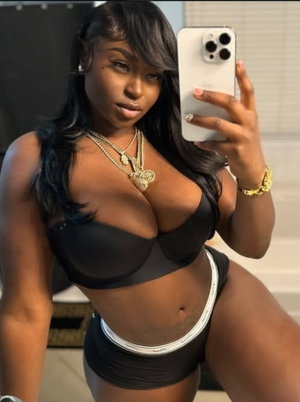 Woman in black bra and shorts taking mirror selfie, wearing gold jewelry, indoor setting.