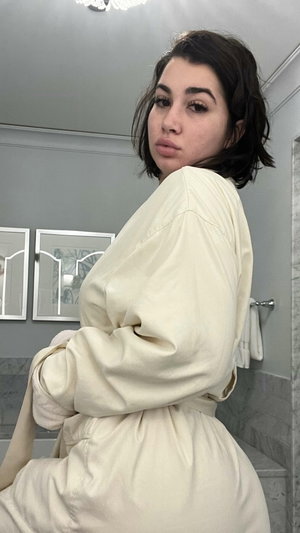 Dark-haired woman in cream robe posing in bathroom.