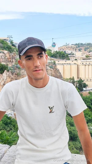 Man in white t-shirt and cap, standing outdoors with rocky hills and buildings in background.