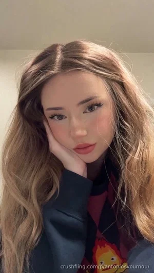 Woman with long wavy hair, wearing a graphic sweatshirt, resting her face on her hand indoors.