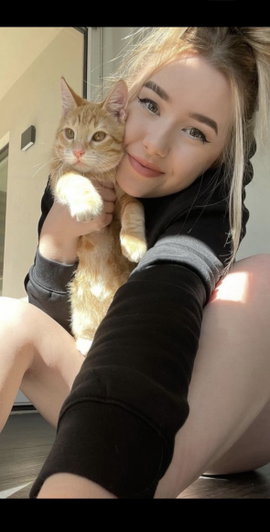 Blonde woman holding orange cat taking indoor selfie.
