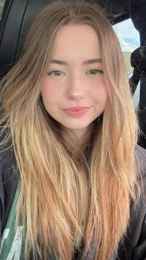 Woman with long blonde hair sitting in a car, wearing a jacket and green shirt.