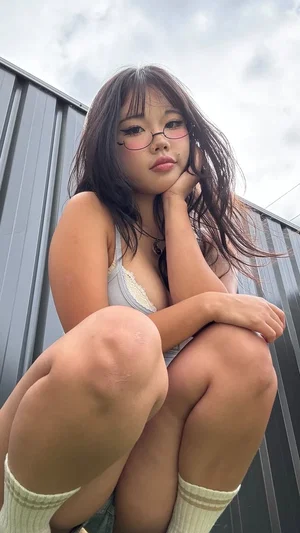 Woman with dark hair and glasses in a gray top and socks, sitting outdoors against a metal wall.