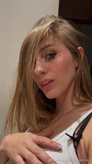 Woman with blonde hair and makeup, wearing a white top with black straps, hand on chest, indoors.