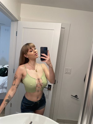 Woman in green crop top and denim shorts takes mirror selfie in bathroom.